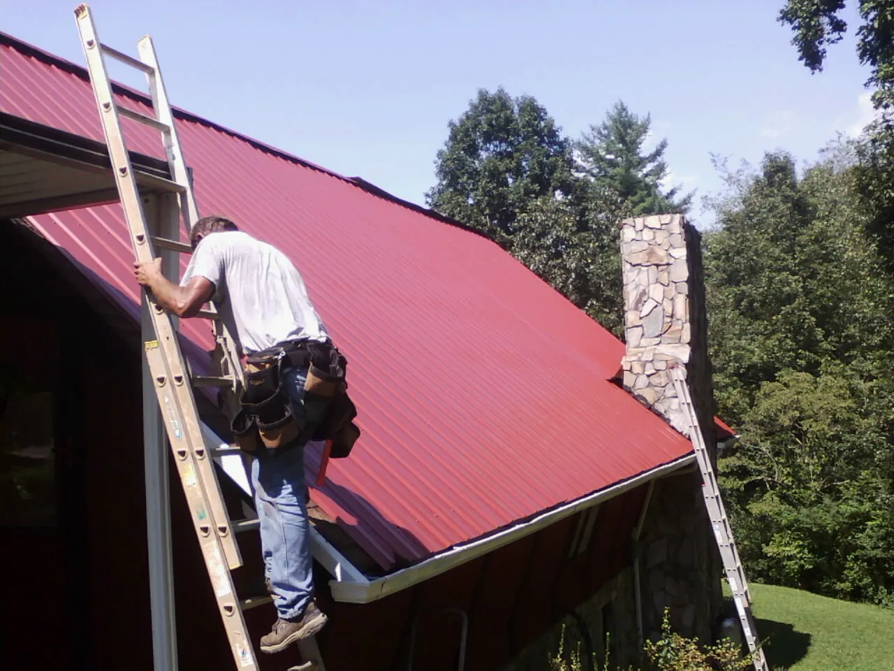 Roofer on a ladder beside a red metal roof and stone chimney — Soffit & Fascia Repair by Roofing Brothers in Shipshewana